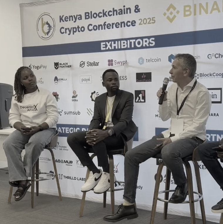 Three panellists on bar stools in front of the Kenya Blockchain & Crypto Conference 2025 exhibitor backdrop; Mik Ruberl, at right, is speaking into a handheld microphone.